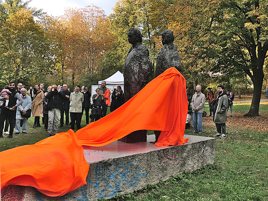 Orangefarbenes Tuch hängt über Statue mit 2 Figuren, dahinter Zuschauende