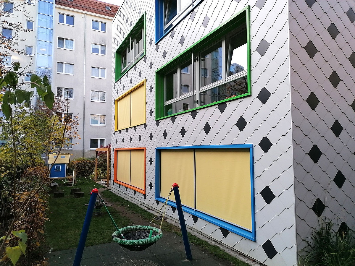 Fassade mit weiß-schwarzem Schieferplattenmuster, farbige Fensterrahmen, schmale Spielfläche, Plattenbauten