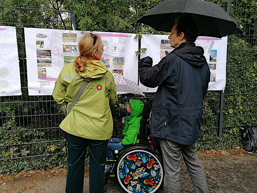 Frau, Mann und Kind im Rollstuhl vor Plakatwand am Zaun mit Regenschirm