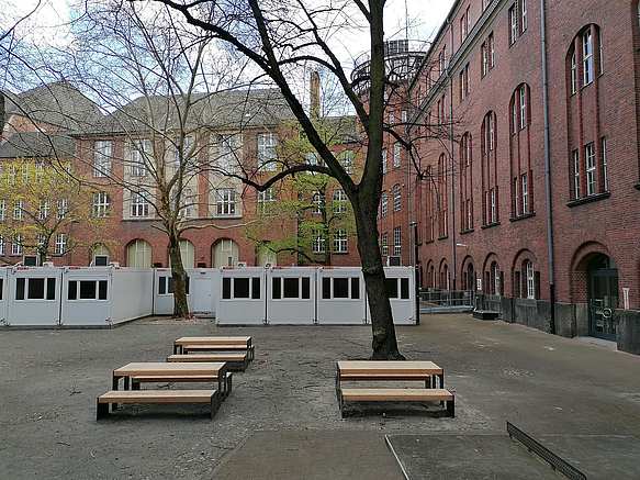 Schulhof mit Tisch-Bank-Kombinationen und Containern, Backsteingebäude, Bäume im Frühjahr