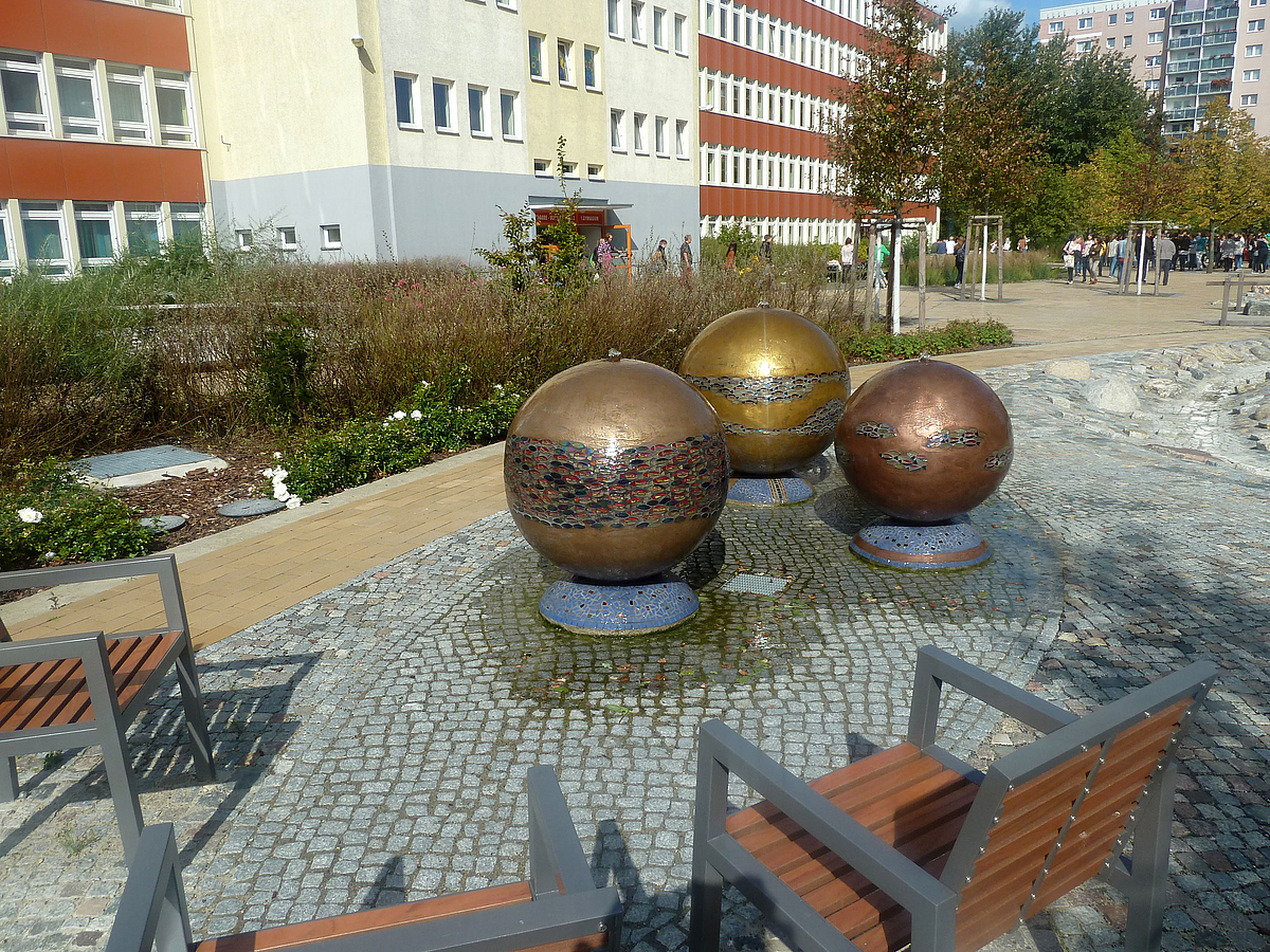 3 Metallkugeln mit Email-Bändern auf gepflasterter Erhebung, 2 Stühle, im Hintergrund das Schulgebäude (Typenbau)
