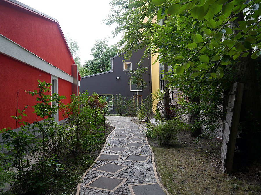 Pflasterweg mit Mosaik vorbei an roter Fassade Eingeschosser auf graues, abgestuftes, modernes Gebäude zu, Grün