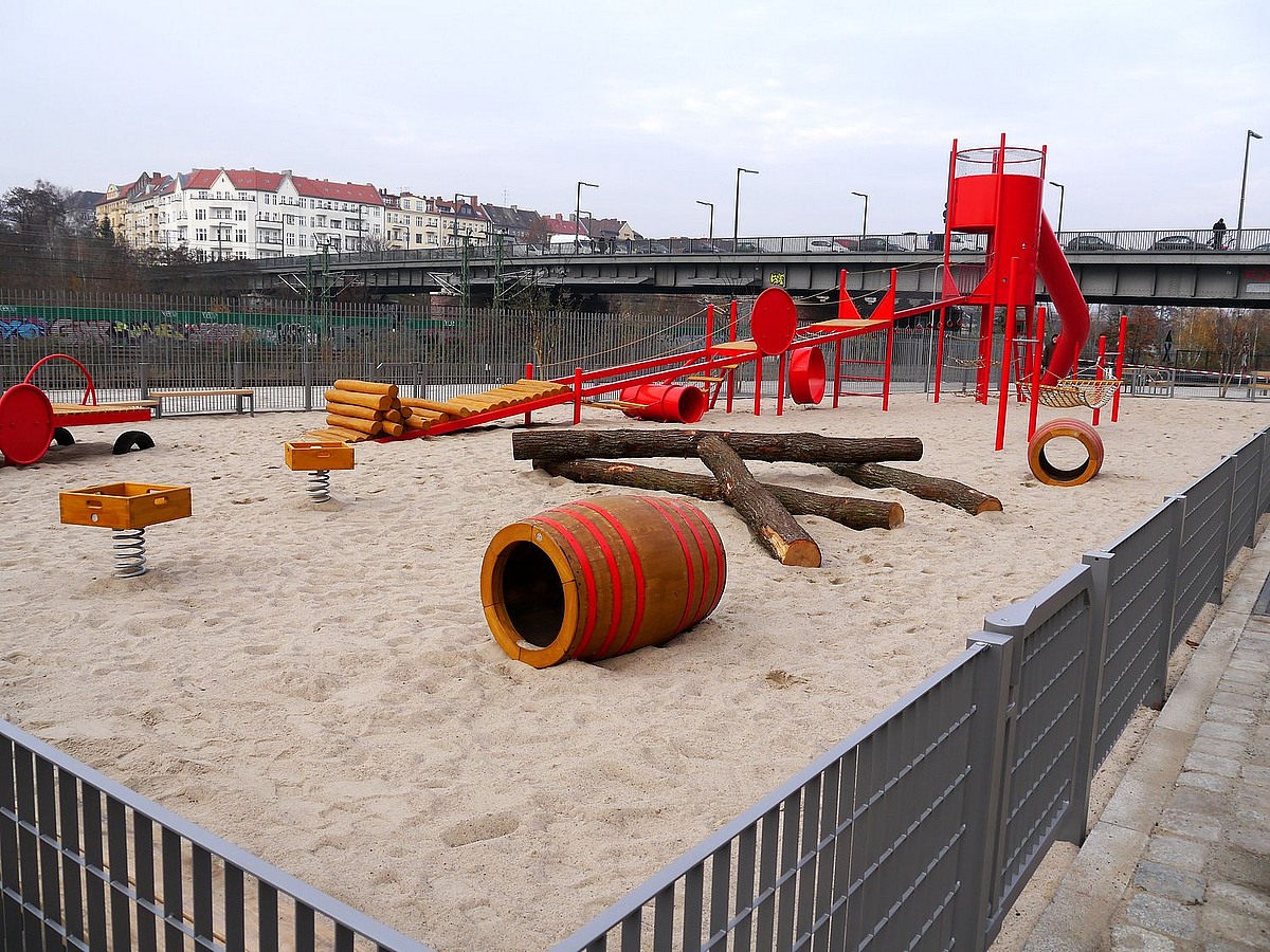 Sandfläche mit rotem Spielgerät aus Metall mit Rampe und Rutschenturm, im Vordergrund kleinere Geräte