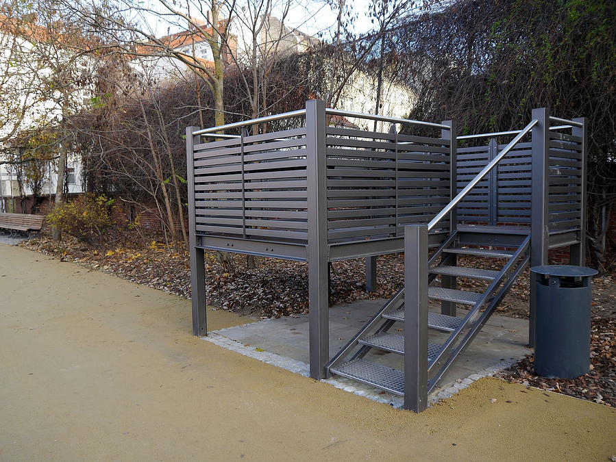 Beigefarbener Weg mit kleinem "Hochsitz" aus Metall mit Treppen inkl. Geländer