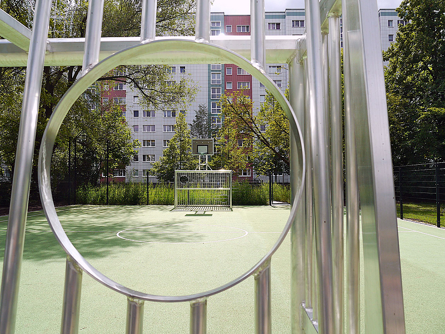 Blick durch Torwand-Loch auf Bolzplatz mit grünem Kunststoffbelag, dahinter Plattenbauten