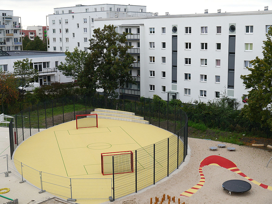 Vogelperspektive auf linsenförmiges, gelbes Ballspielfeld mit roten Toren und Ballfangzaun