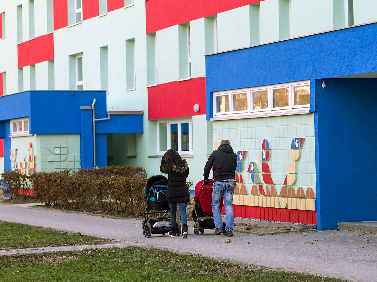 Frau und Mann jeweils mit Kinderwagen vor hellblauem Gebäude mit roten und blauen Akzenten