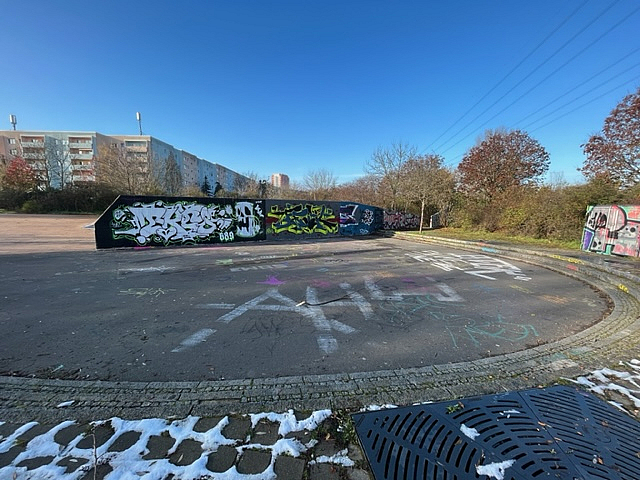 Ovale Asphaltfläche, mittig Mauer mit Graffiti, Wohngebiet im Hintergrund
