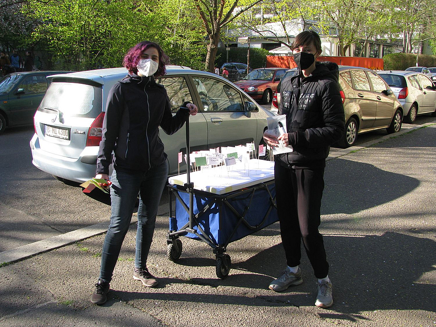 2 Personen mit Maske und Handwagen mit Material auf Gehweg