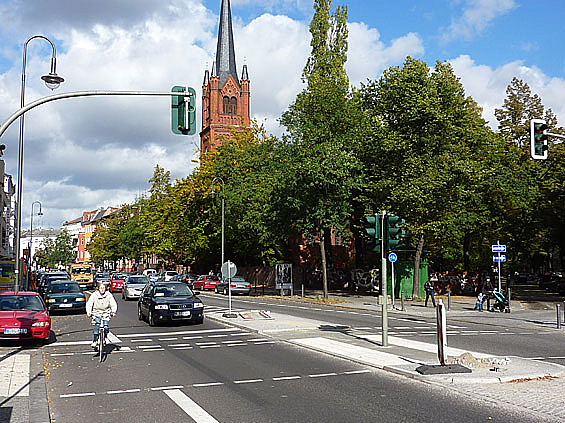 Straße mit Fußgängerinsel, Radstreifen und Parkszone, im Hintergrund Kirche und Grünanlage