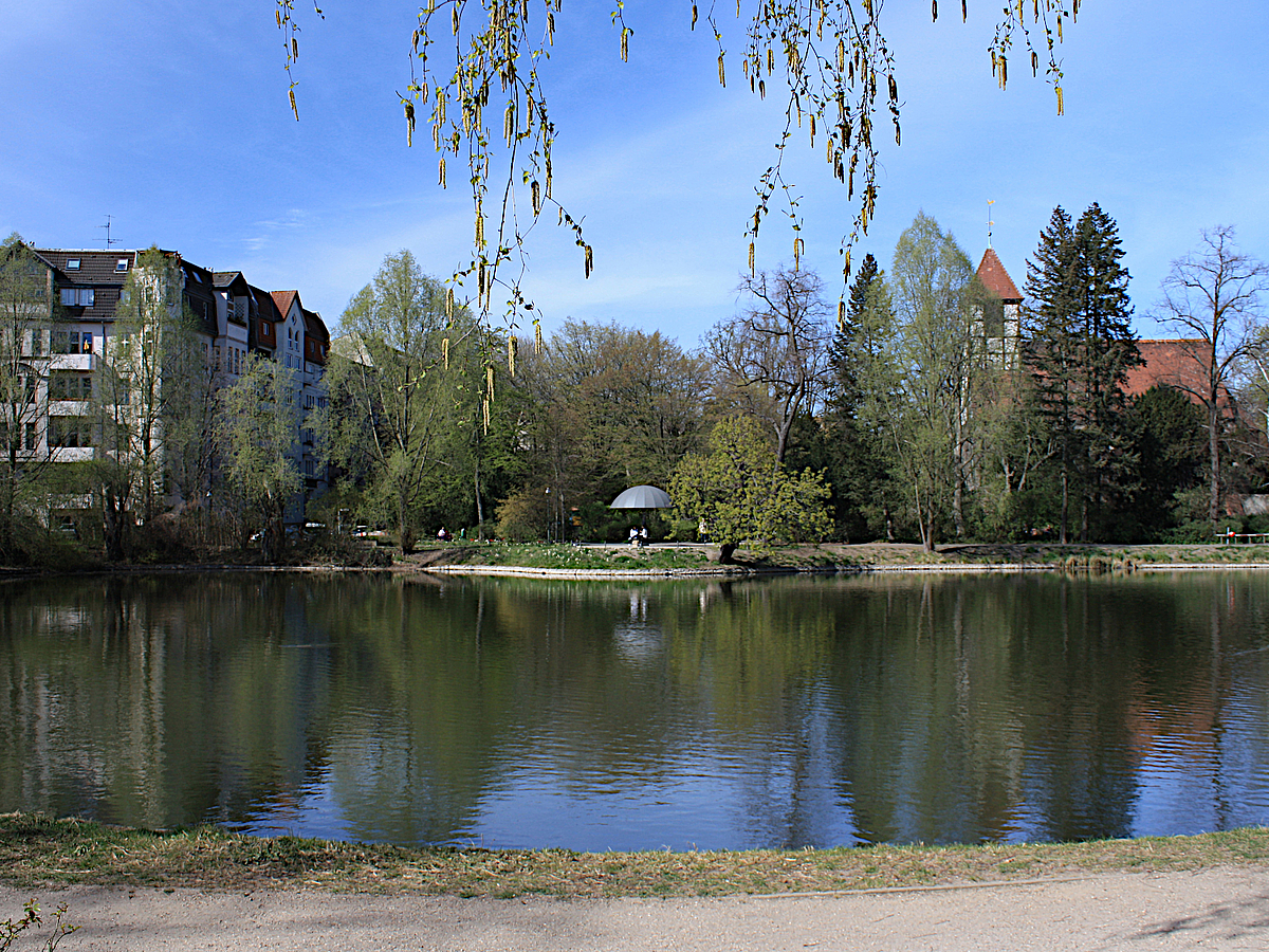 Blick über Teich zu pilzförmigem Pavillon, Wohnhaus, Kirche im Frühling