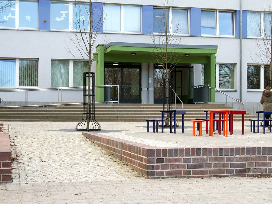 Gepflasterter Hof mit farbigen Tischen und Schulgebäude im Hintergrund