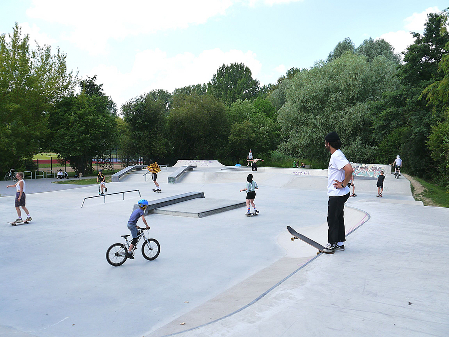 Betonfläche mit radfahrendem Kind und anderen Sportlern, Skateboarder wartet am Rand
