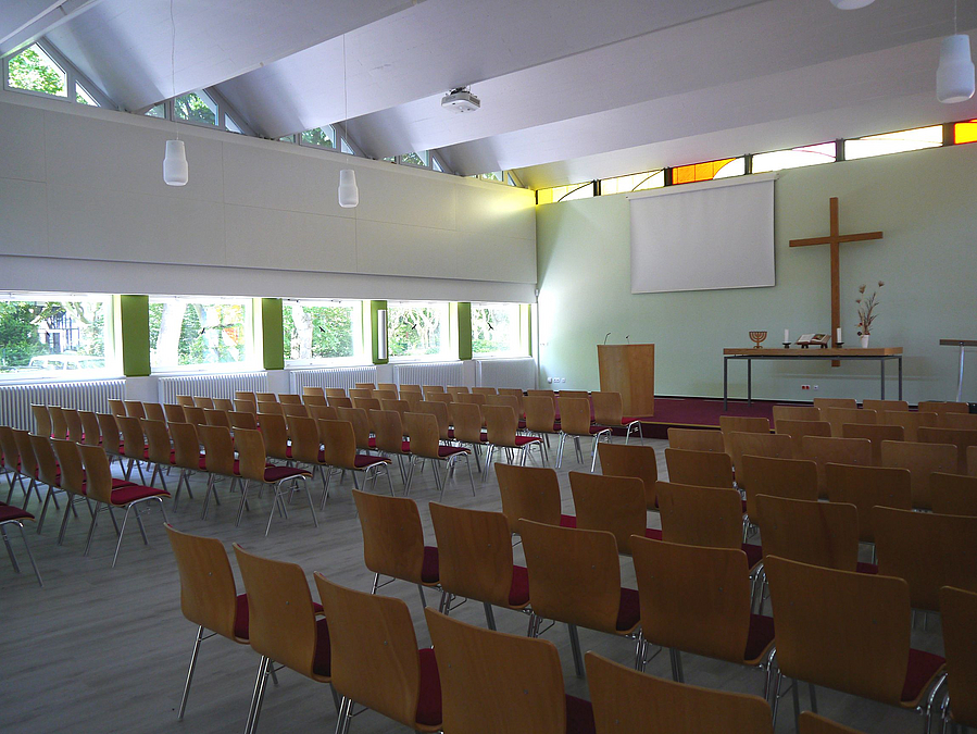 Saal mit Faltendach, Fensterband, Stuhlreihen, Kreuz und großem Bildschirm an der Wand
