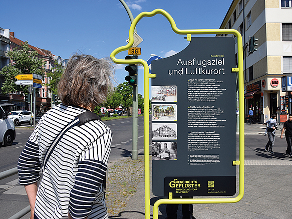 Frau betracthet Infostele auf der Straße mit gelbem Metallrahmen in Form eines Hutes und schwarzer Tafel zum Thema Ausflugs-Lokal