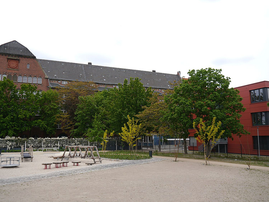 Blick über Sandfläche auf Spielplatz, Altbauschule und rote, moderne Fassade