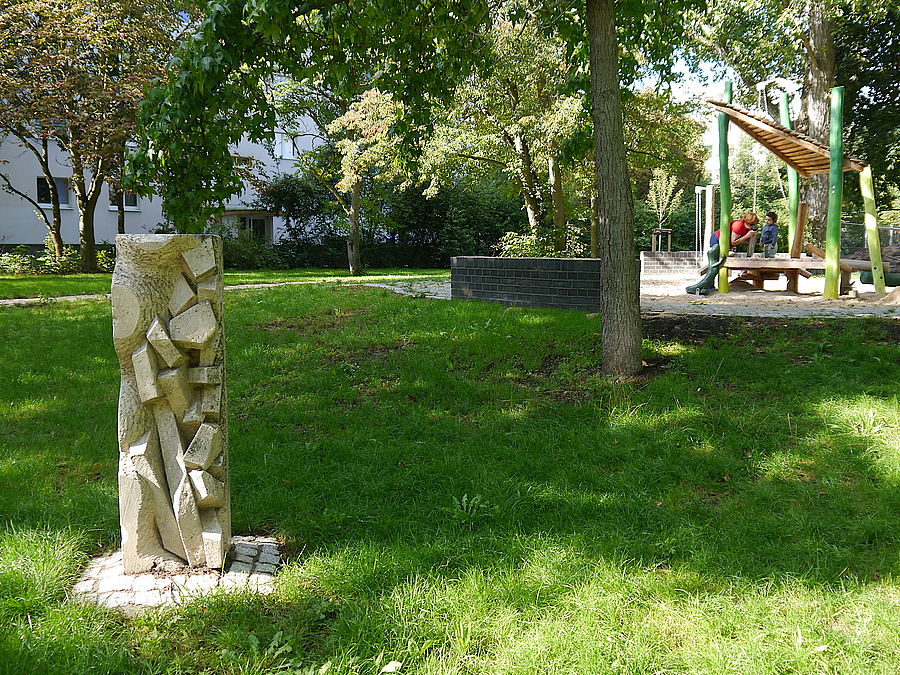 Niedrige Stele mit Relief auf Rasen, im Hintergrund Spielplatz