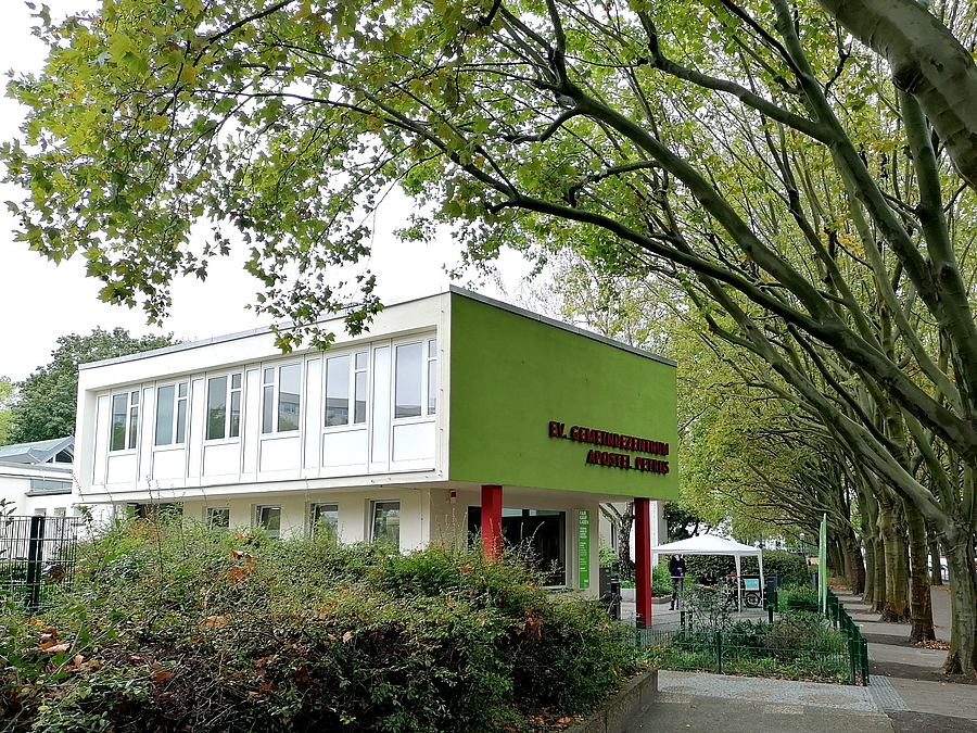 Vorspringes Obergeschoss mit grüner Front auf roten Pfeilern an Baumallee, davor Grün
