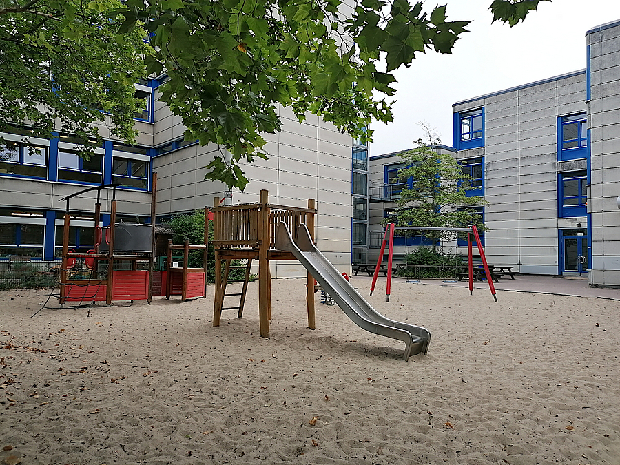 Helle Sehulgebäude, Sandfläche mit Rutsche und mehr, Laubbaum