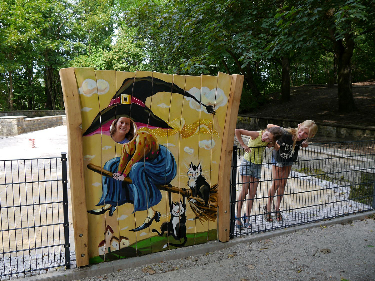 Holzwand mit Hexe auf dem Besen und Guckloch mit Kind, 2 Mädchen schauen von innen über den niedrigen Zaun auf das Bild