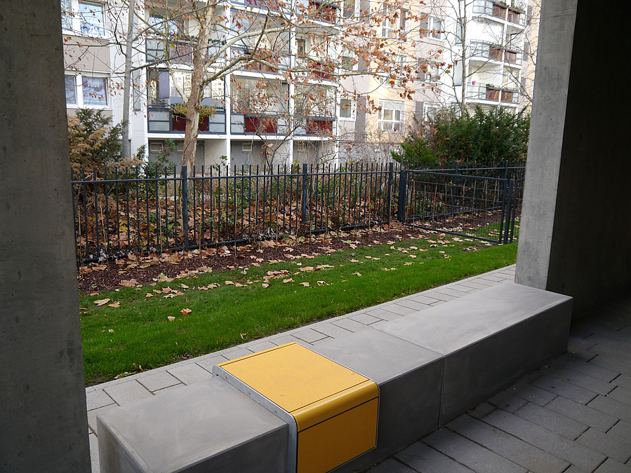 Betonrahen mit gelber Sitzfläche, dahinter Wiese, Zaun, Plattenbau