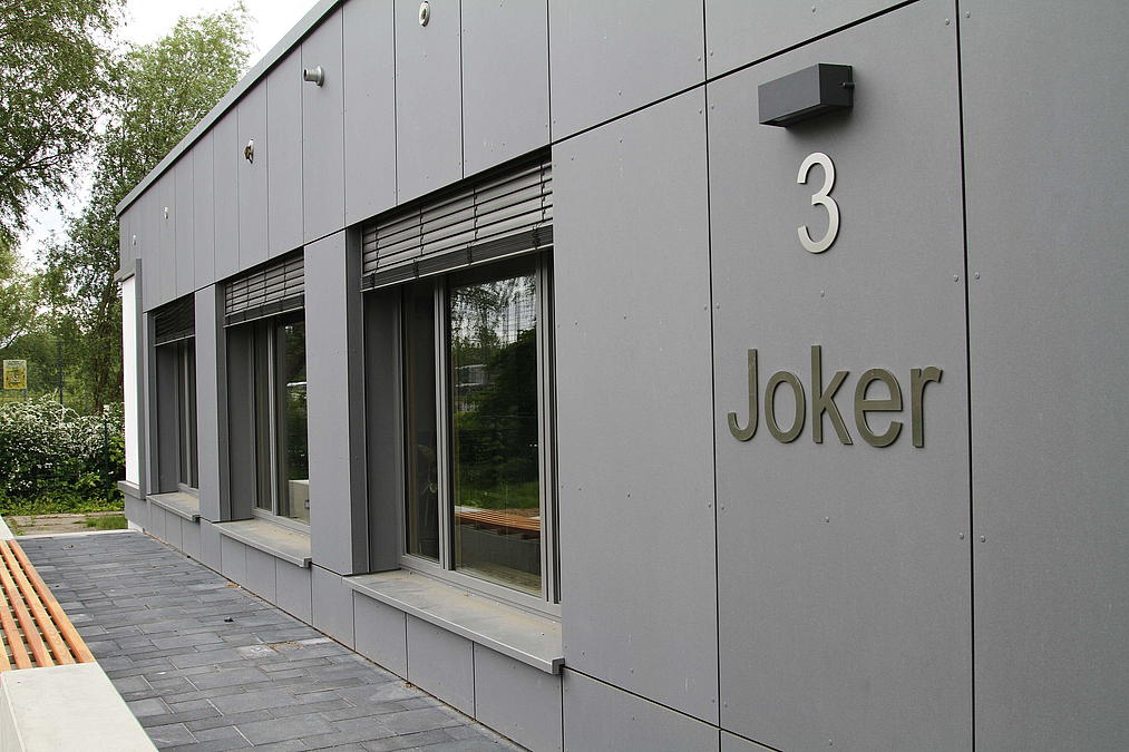 Graue Fassade mit bodentiefen Fenstern und Schriftzug "Joker"