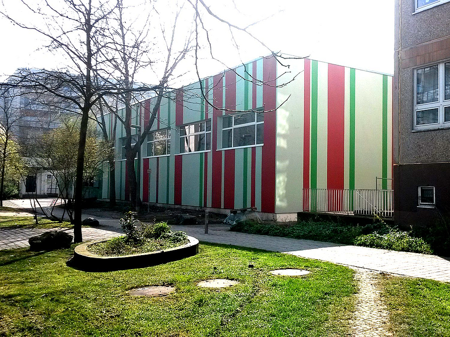 Fassade mit beigen, roten und grünen Streifen, davor Grünfläche