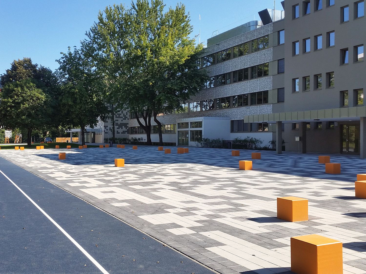 Laufbahn im Anschmitt, Hof mit orangen Kuben zum Sitzen auf QR-Code-Platten-Muster, daniertes Schulgebäude