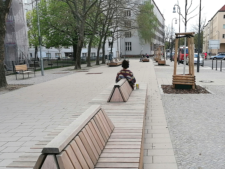 Lange Holzdecks mit niedriger Lehne auf breiter Promenade, eine Person sitzt an der Schmalseite