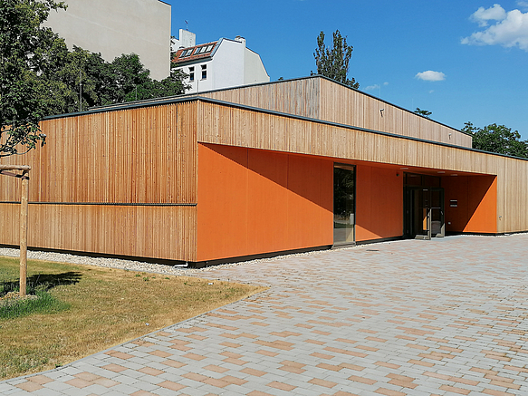 Eingeschossiges Gebäude mit Holzlamellen-Fassade und orangefarbener Eingangsfront
