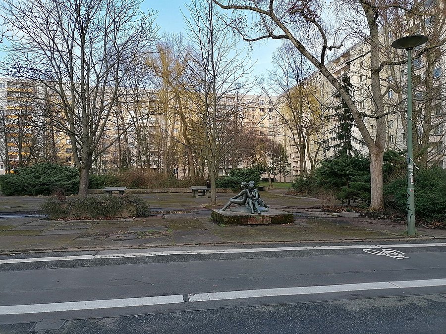 Blick über Asphalt-Straße mit Radweg auf Skulptur, kahle Bäume auf befestigtem Platz