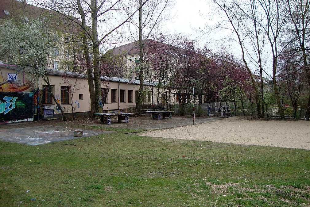 Rasen, Sandfläche, 2 Tischtennisplatten, im Hintergrund heller, langgezogener Flachbau 