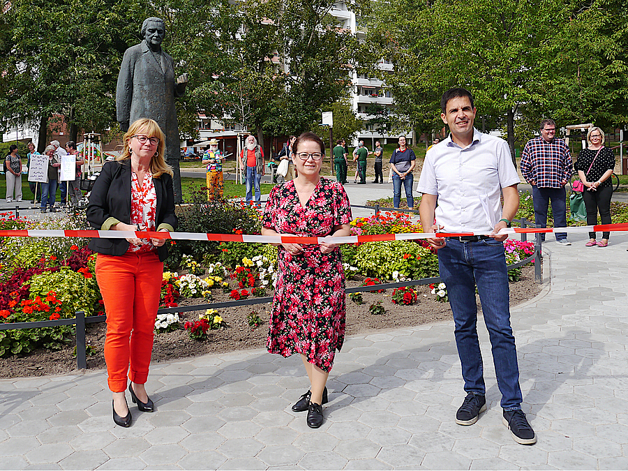 3 Personen hinter Flatterband, dahinter überlebensgroßes Denkmal im Blumenbeet