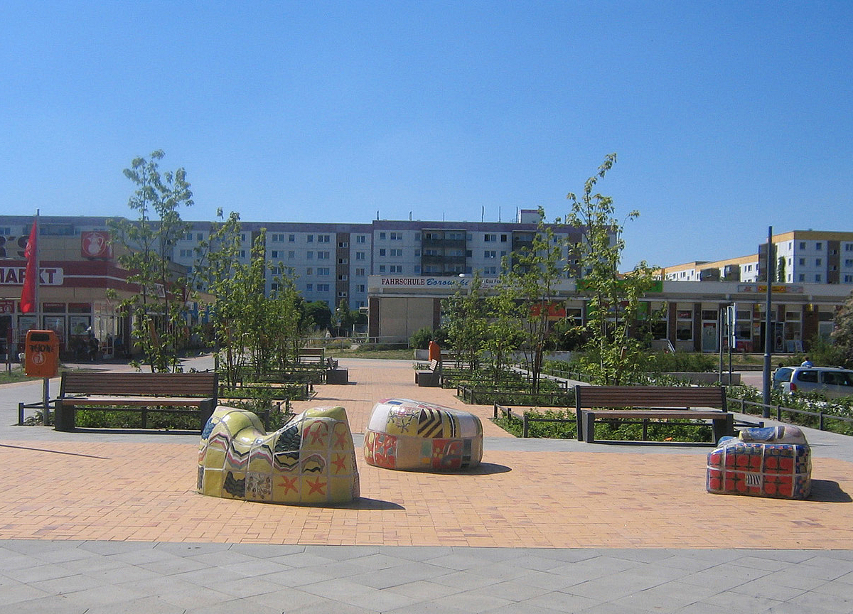 Platz mit Kusntobjekten aus Keramik zum Sitzen, Bänke, junge Bäume, im Hintergrund Einkaufszentrum, Wohnbauten