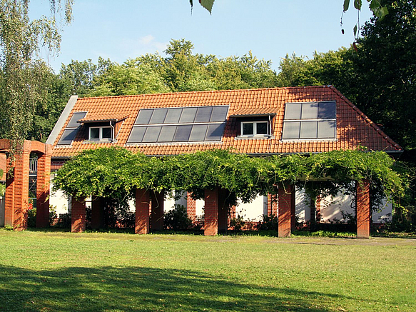 Klinkerbau mit großen Dachgeschossfenstern vor begrünter Pergola, dahinter dichtes Grün