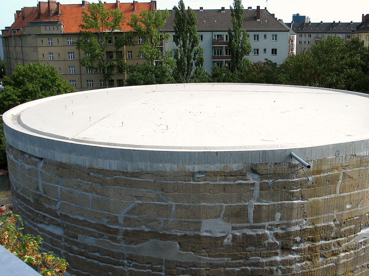 Großer, gemauerter Zylinder mit saniertem Dach, dahinter Dächer der Wohnbebauung