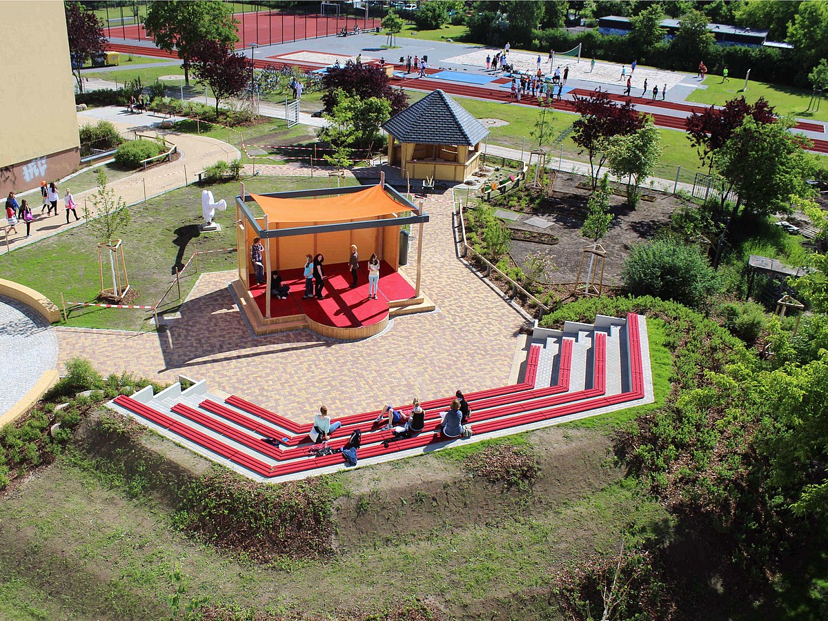 Vogelperspektive mit Bühne und Amphithater in orange und rot im Zentrum, Pavillon, Sportanlagen und Pflanzungen