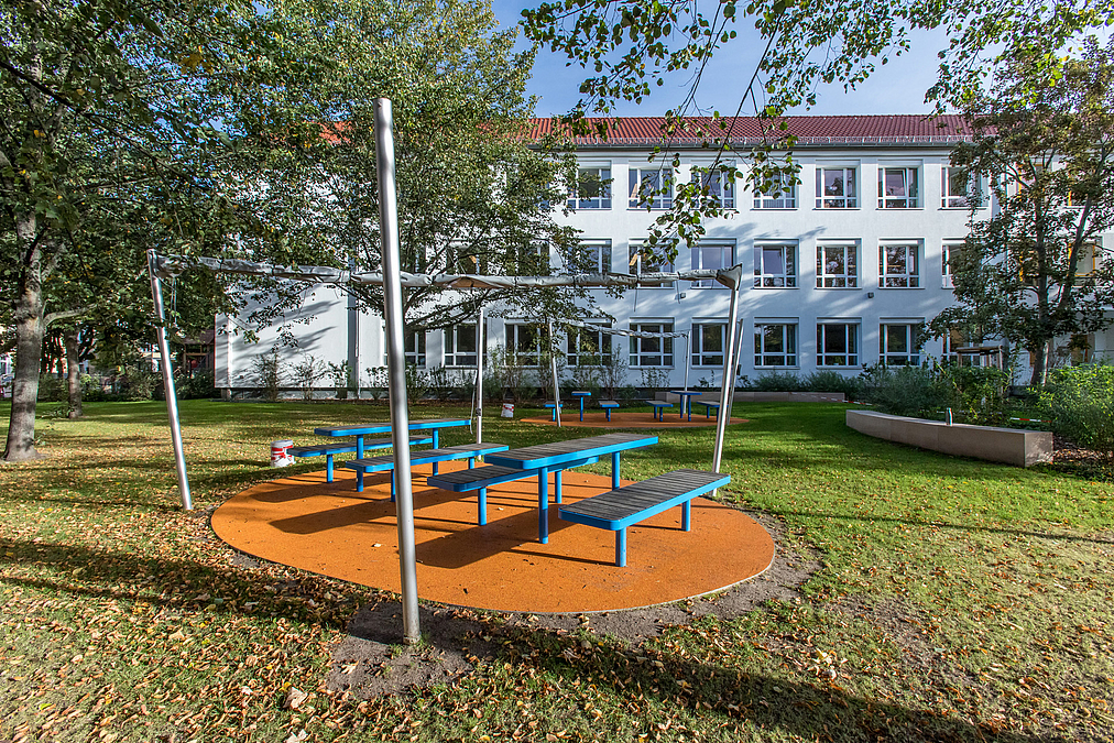 Ovale, hellrote Kunststofffläche in Rasenfläche mit Tischen und Bänken in Blau, Laubbäume, helles, älteres Schulgebäude im Hintergrund