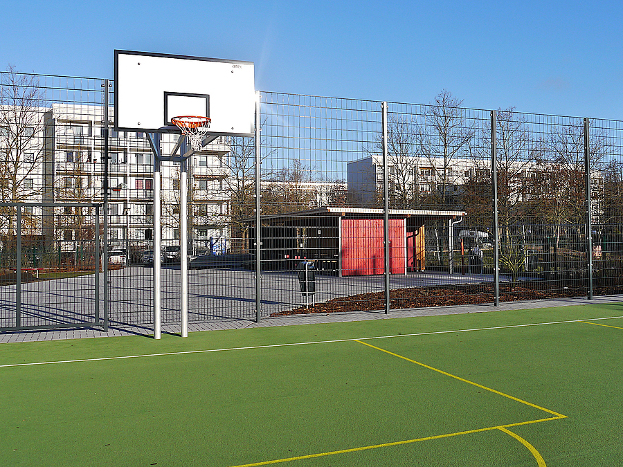 Grünes Sportfeld mit Linierung, Basketballkorb, dahinter rotes Gebäude