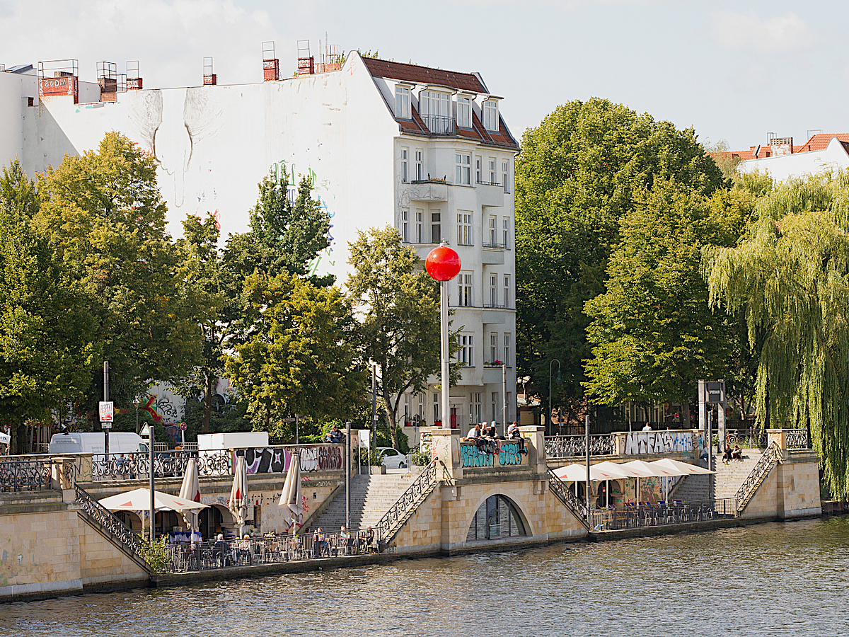 Blick über Fluss auf Ufermauer mit Terrasse, Mast und rotem Ballon