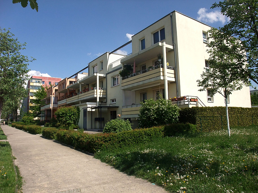 Blick auf weißen Dreigeschosser mit Balkonen und kleine Mietergärten
