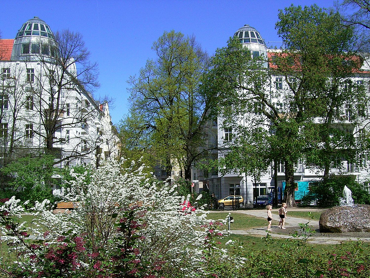 Platz mit blühenden Büschen und sanierter Gründerzeit-Bebauung im Hintergrund