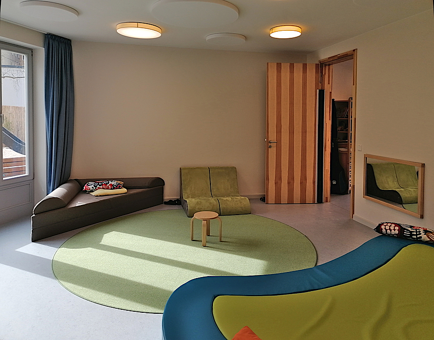 Raum mit grünem Teppich, buntem Liegesofa, weiteren, niedrigen Sofas