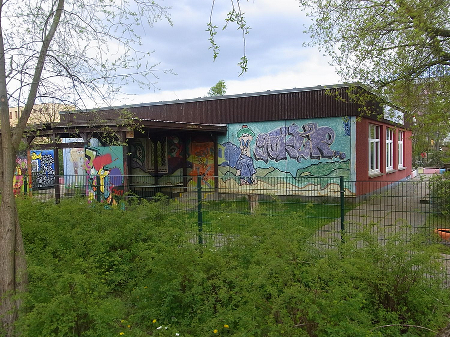 Flachbau mit gestaltetem Graffiti, davor Zaun und Unkraut