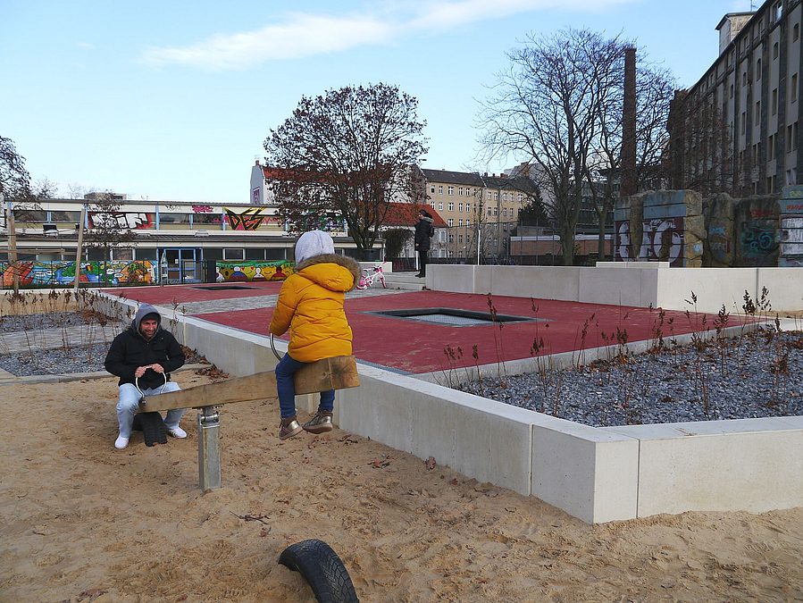 Wippe mit Mann und Kind auf Sandfläche, in Beton eingefasste Terrasse mit Pflanzung auf Schotter und Trampolin in rotem Kunststoff