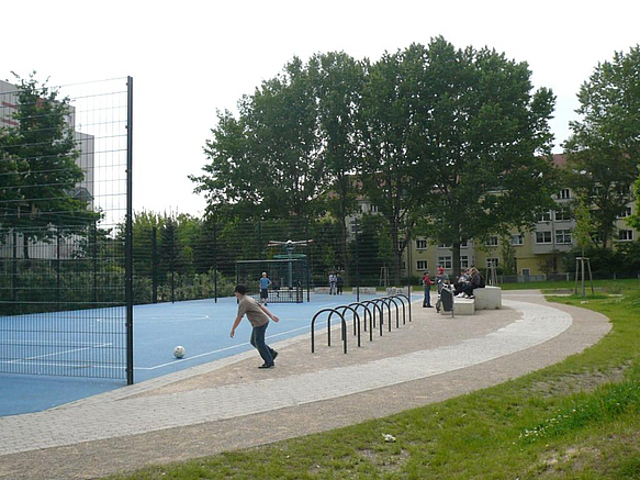 Linsenförmige Fläche mit blauem Bolzplatz, Fahrradbügeln, Quader zum Sitzen, Bäume, Wohnhäuser