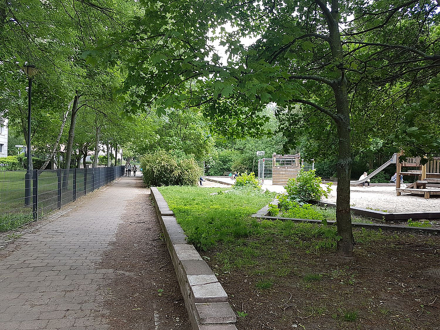 Von Bäumen gesäumter Fußweg, rechts Spielplatz, links Zaun