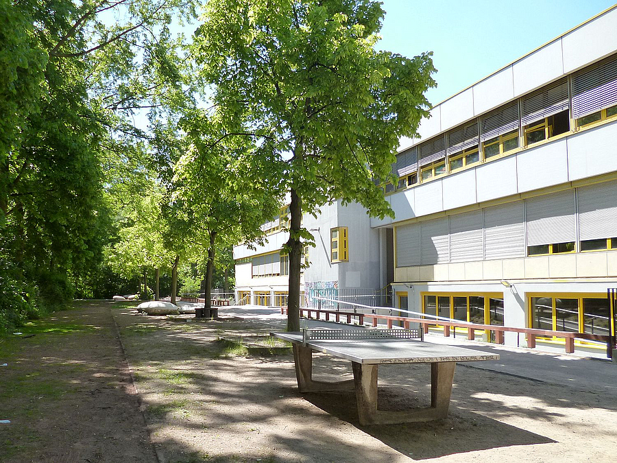 Zweistöckiges Schulgebäude, davor Tischtennisplatten und Bäume, unbefestigter Boden