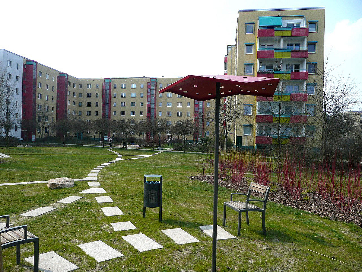 Weg, eiserner Sonnenschirm und Bänke auf einer Grünfläche zwischen sanierten Plattenbauten