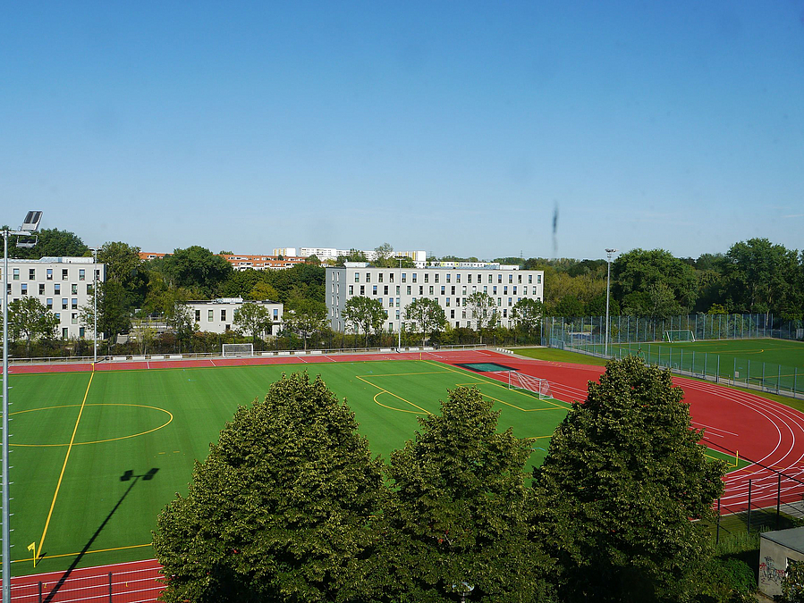 Blick über Bäume auf Stadionkurve und halbes, grünes Fußballfeld, im Hintergrund Gemeinschaftsunterkunft